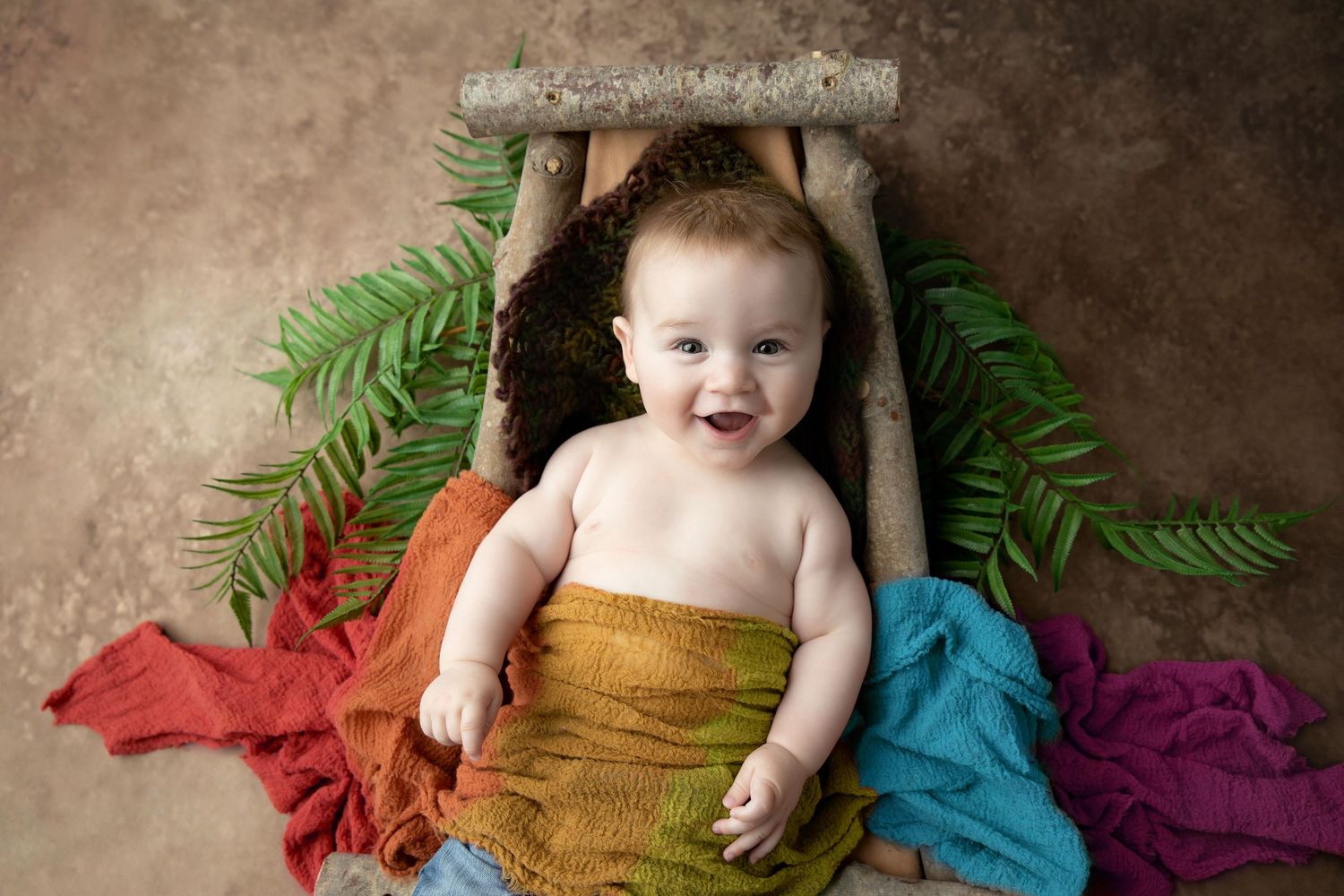 Smiling baby lying in a wooden prop bed with rainbow wrap and green fern leaves in a studio portrait. Family Photographer Cork.