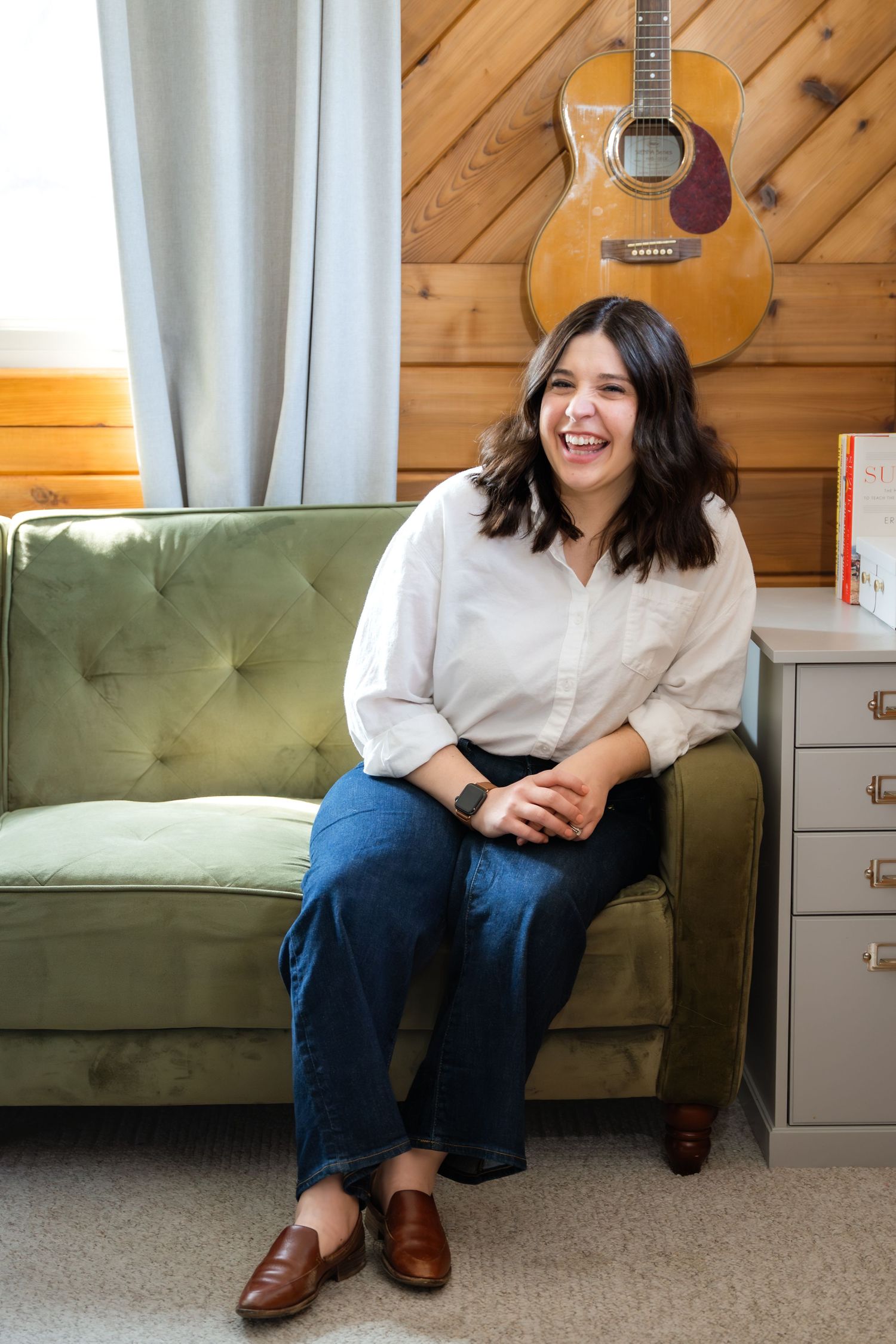 A smiling woman in a white shirt and jeans sits on a green tufted sofa with an acoustic guitar on the wood wall behind her.