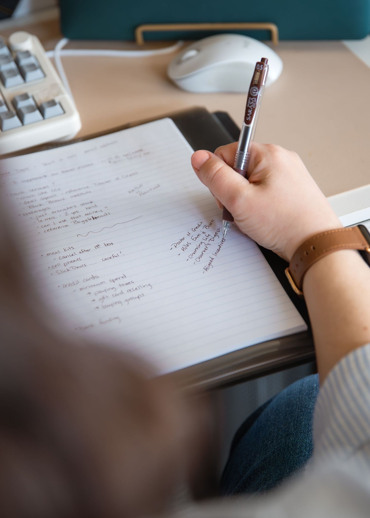 Person writing notes in a lined notebook with a pen, wearing a brown watch, at a desk with a keyboard and mouse.