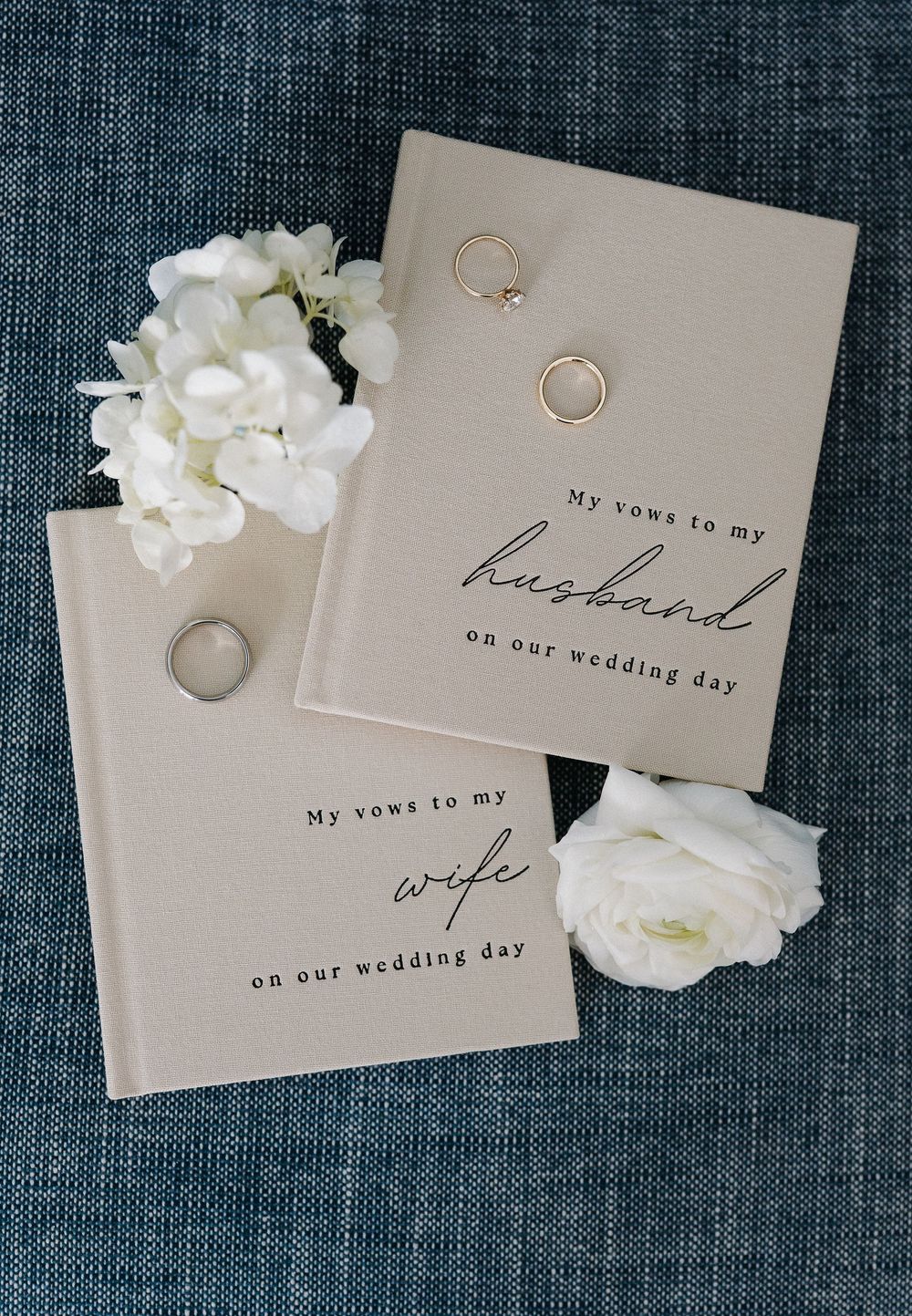 Two elegant wedding vow books for husband and wife displayed with gold rings and white hydrangea flowers on blue linen.