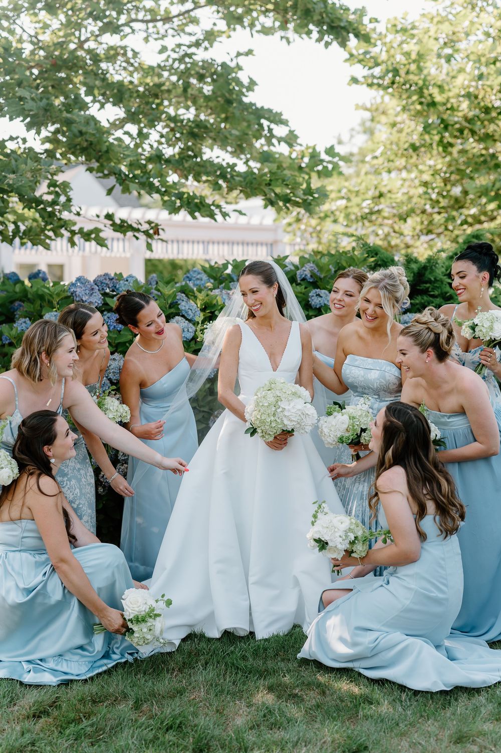 Bride in white gown surrounded by bridesmaids in light blue dresses holding white floral bouquets outdoors.