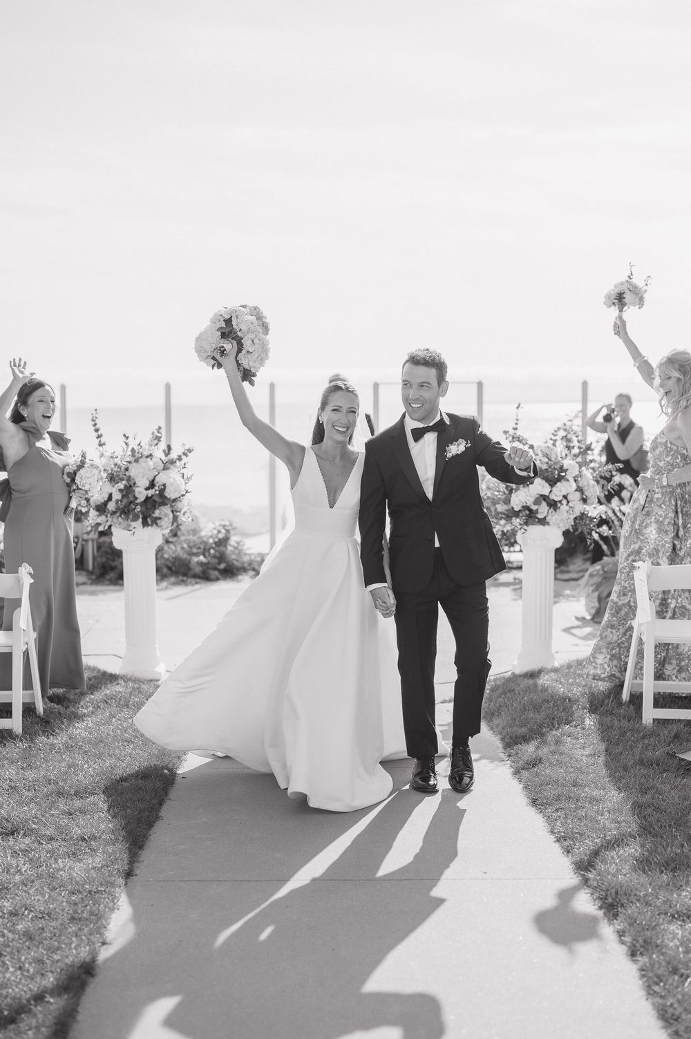 Black and white photo of joyful newlyweds walking back down the aisle after outdoor oceanside wedding ceremony.