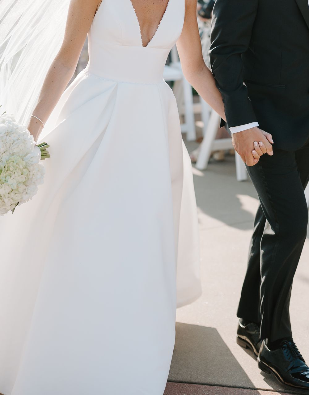 Bride in elegant white V-neck wedding gown holding bouquet, walking hand in hand with groom in black suit.