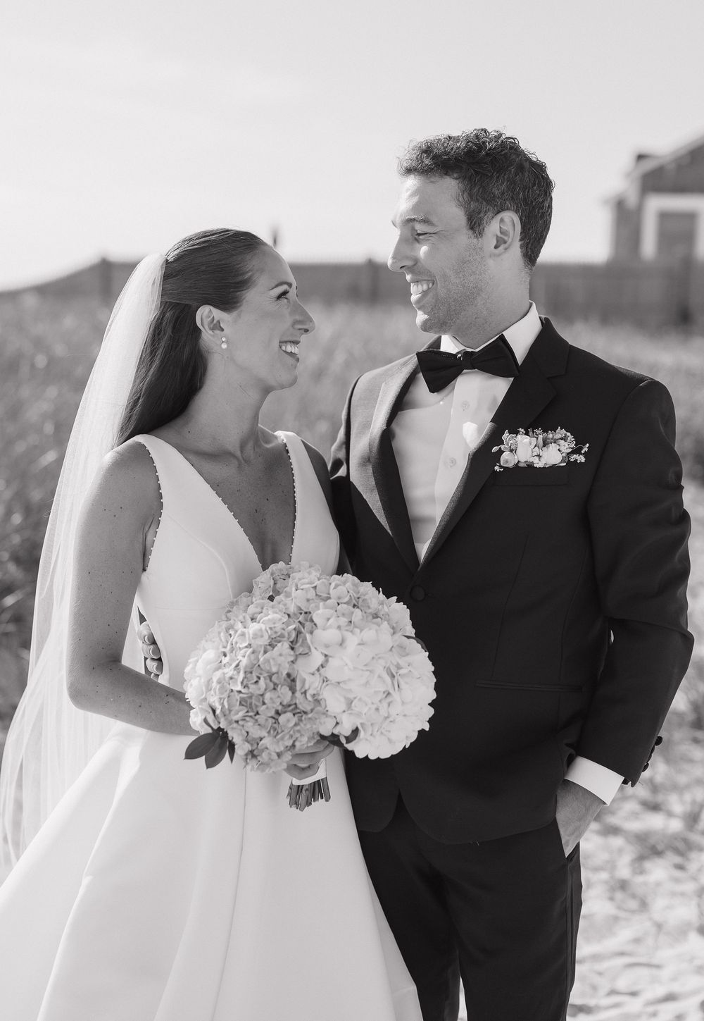 Black and white portrait of smiling bride and groom on beach, bride holding white hydrangea bouquet.