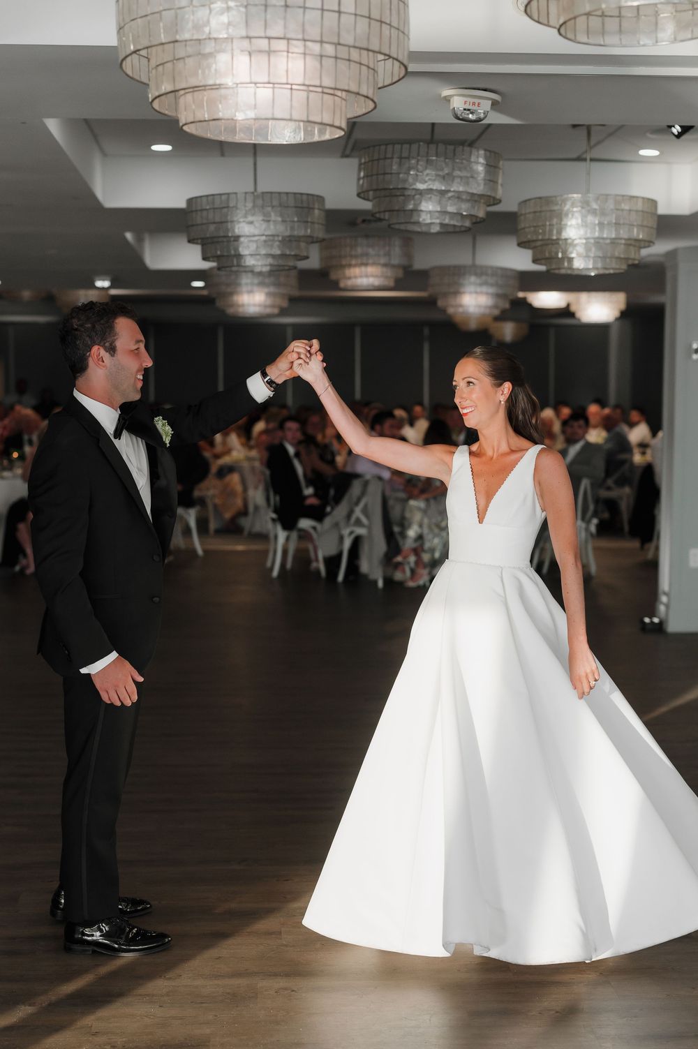 Bride and groom share first dance in elegant ballroom with chandelier lighting at wedding reception.