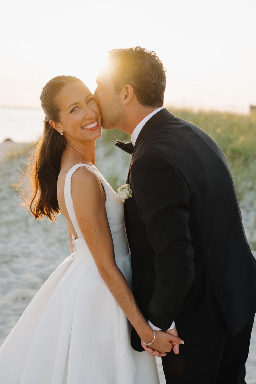 Groom kisses bride on cheek as she smiles on a sandy beach at golden hour during their wedding.