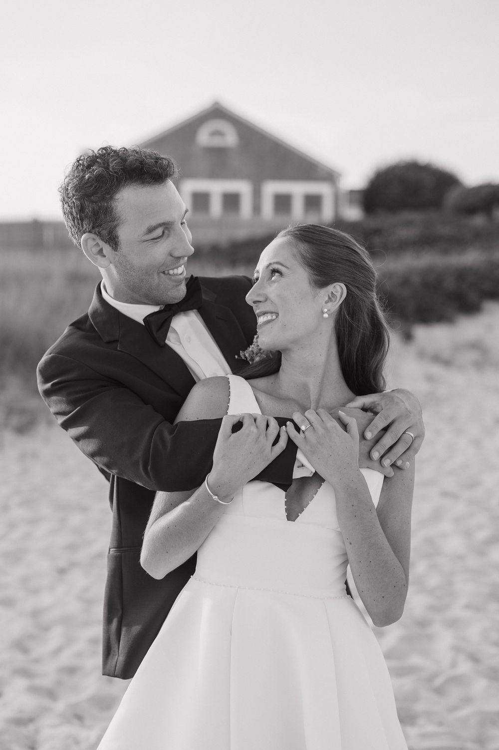 Black and white photo of a smiling bride and groom embracing on a sandy beach with a house in the background.