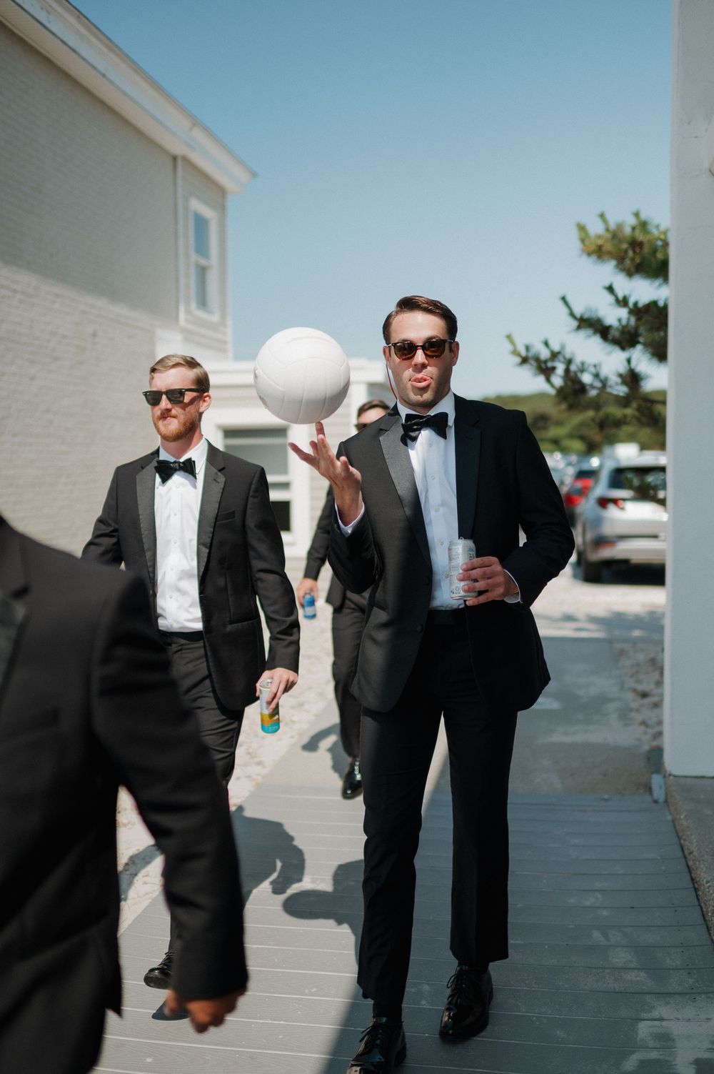 Groomsmen in black tuxedos and bow ties spinning a ball outdoors on sunny wedding day.