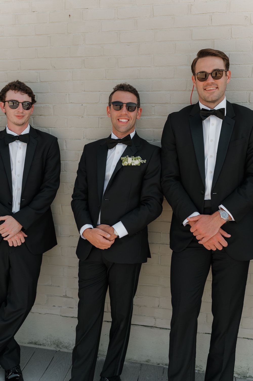 Three groomsmen in black tuxedos and bow ties with sunglasses pose against a white brick wall at a wedding.