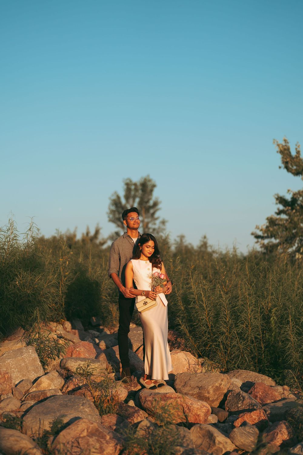 Couple standing on rocks at golden hour, woman in white gown holding flowers, man embracing her with green shrubs behind.