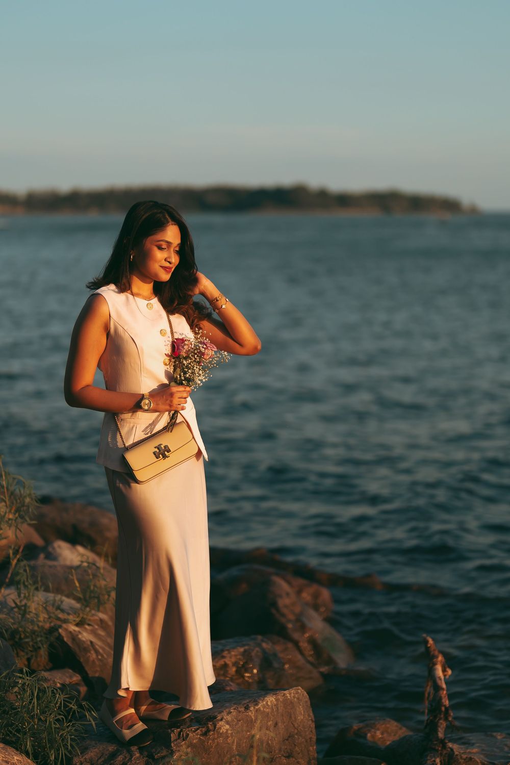 Young woman in white dress holding flowers stands on rocky shoreline at golden hour with ocean backdrop.