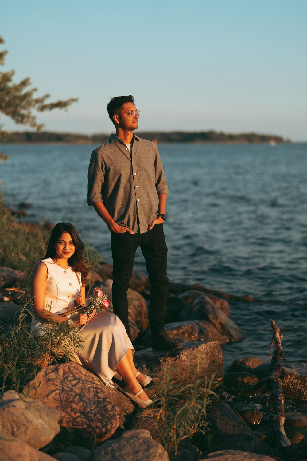 Couple posing by rocky lakeside at golden hour, woman sitting with flowers, man standing in gray shirt and black pants.