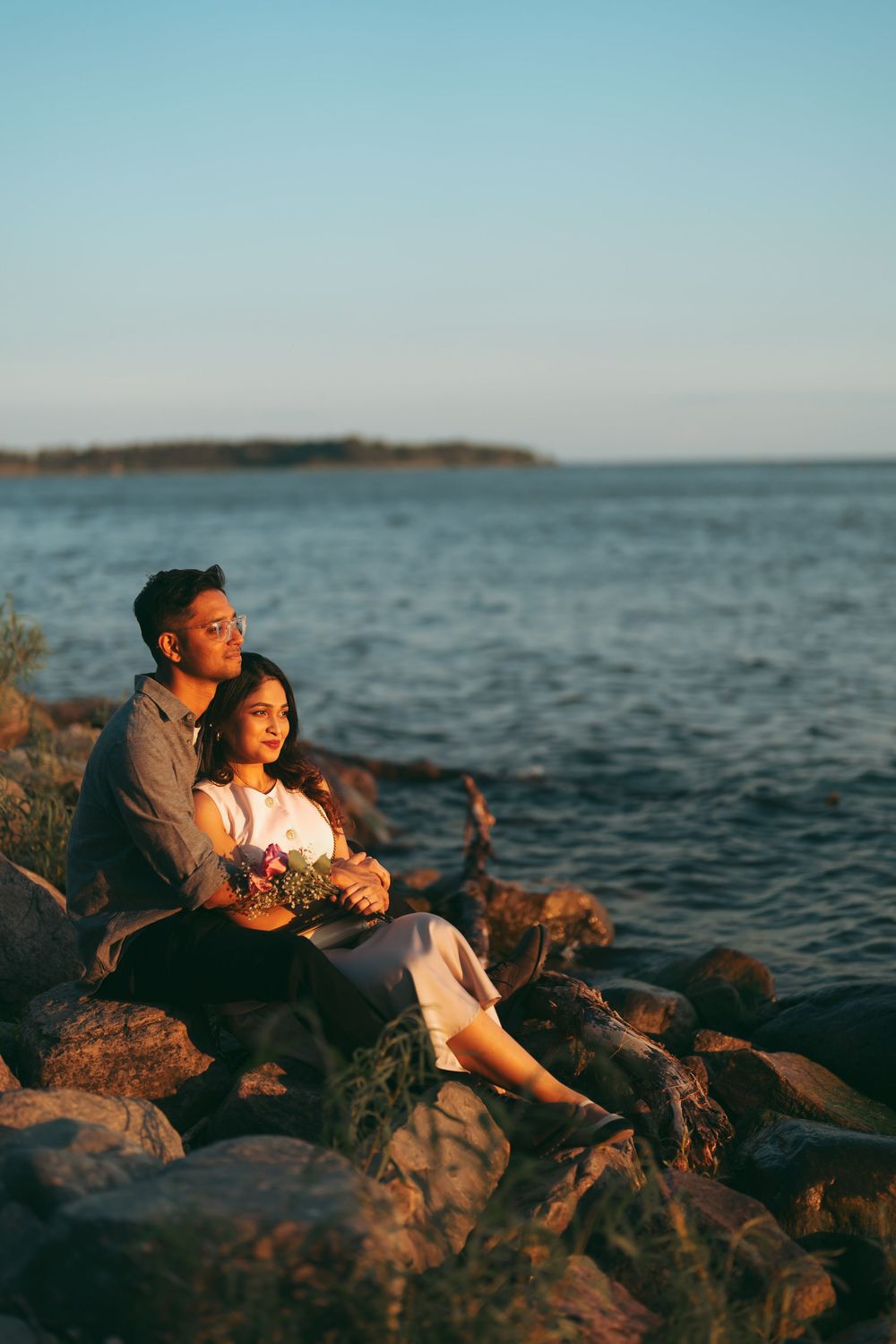 Couple sitting on rocky shoreline at sunset, man embracing woman holding flowers, with calm ocean water in background.