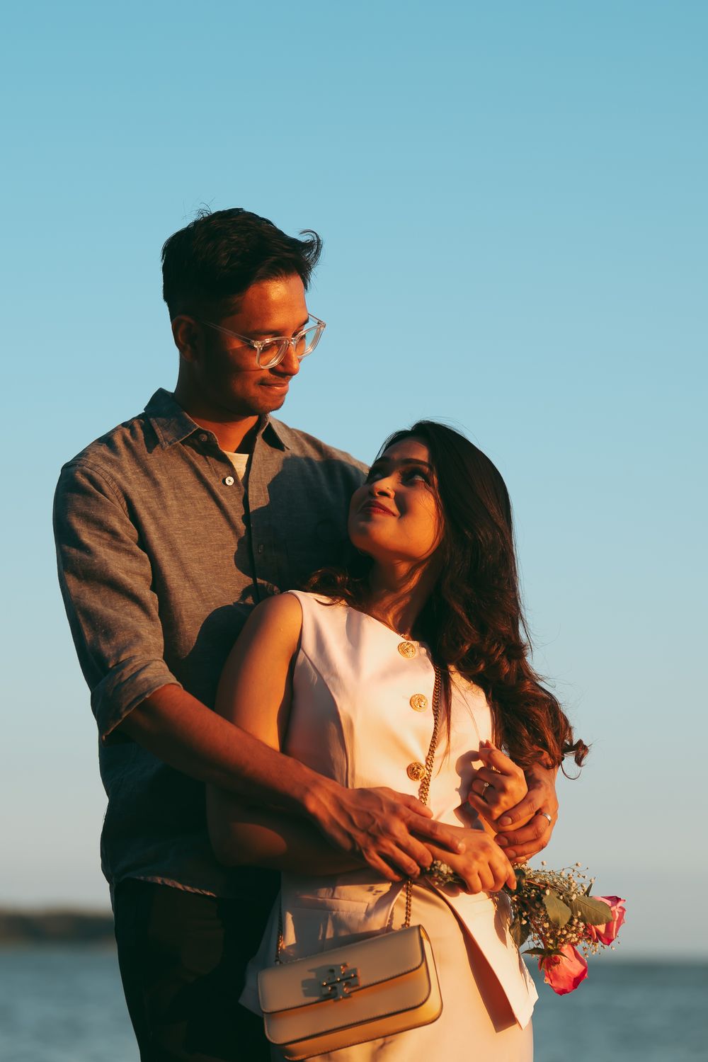 Romantic couple embracing at sunset near the water, woman holding roses, warm golden light.