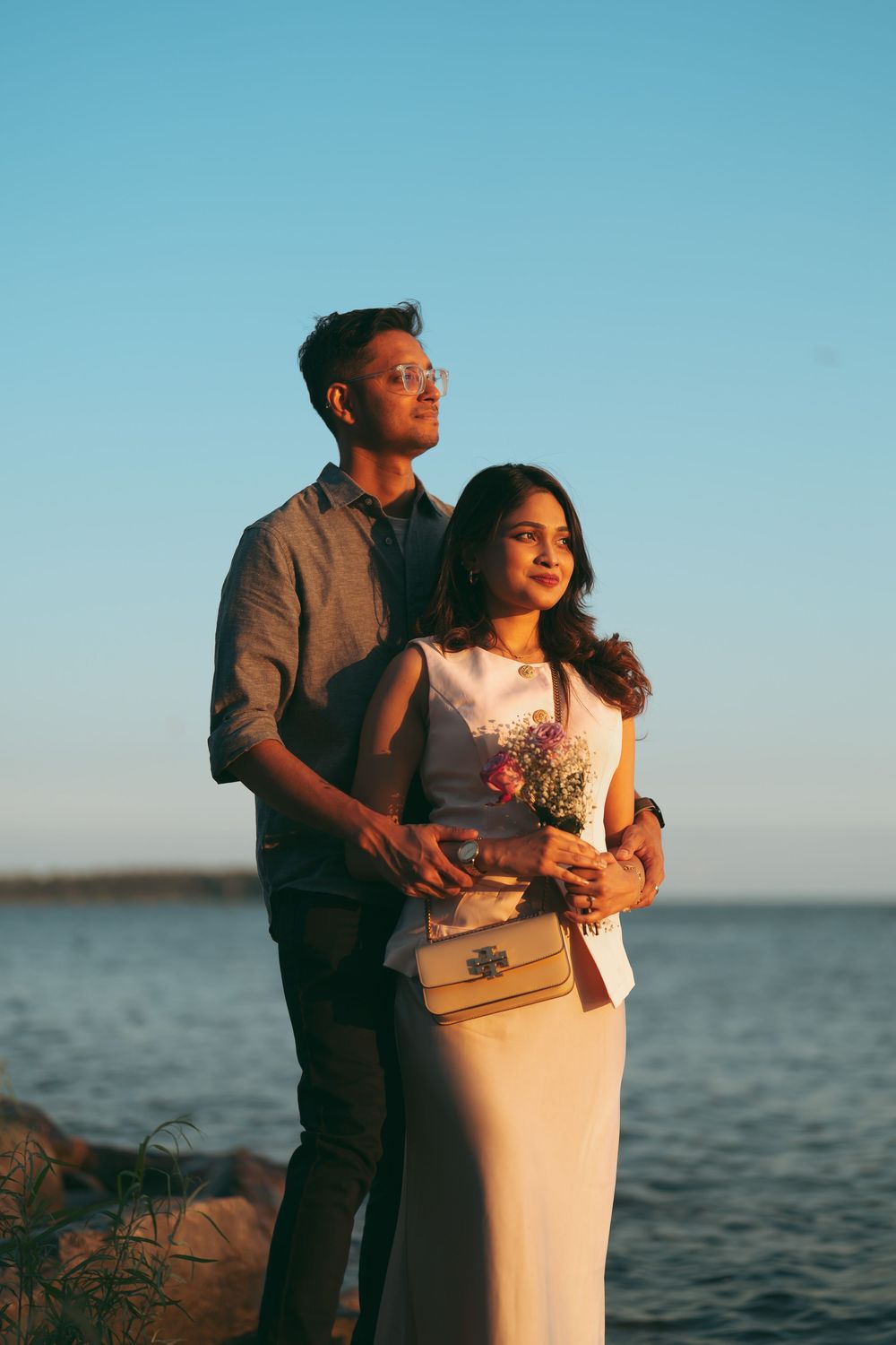 Couple standing by the ocean at sunset, woman holding flowers, man embracing her from behind in warm golden light.