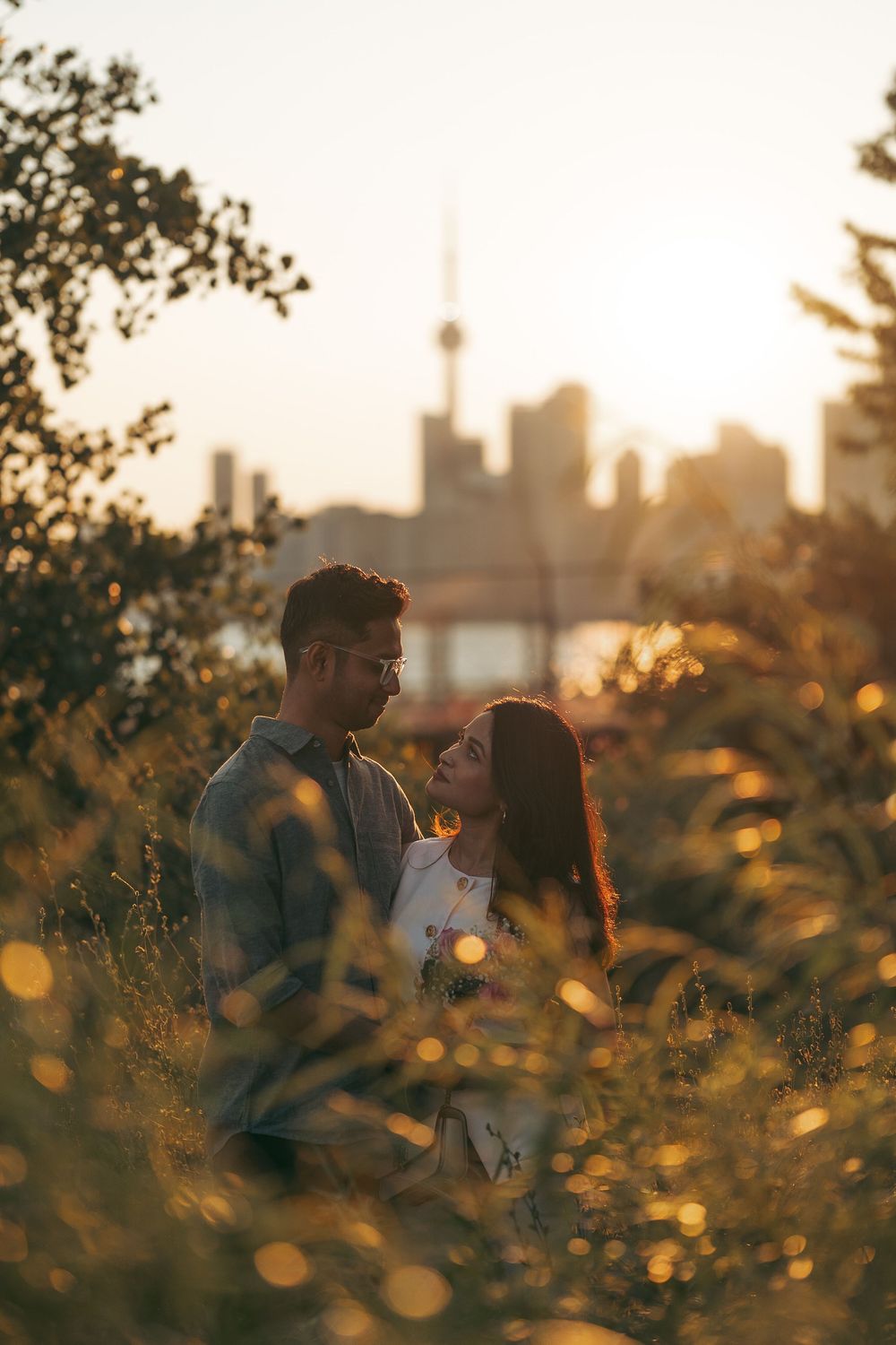 Couple embracing at golden hour with Toronto skyline in background, surrounded by tall grass and warm bokeh lights.