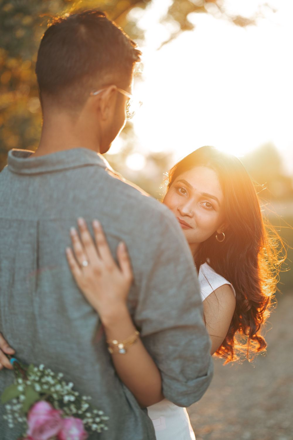 Couple embracing at golden hour sunset, woman with long hair looks back while holding flowers outdoors.