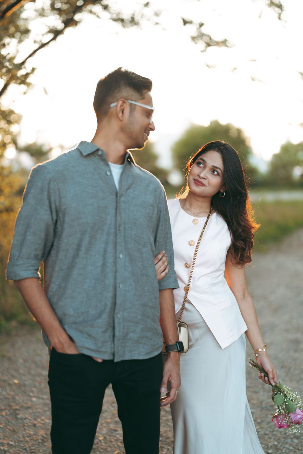 A couple walks together on a scenic path at golden hour, the woman holding flowers while gazing lovingly at her partner.