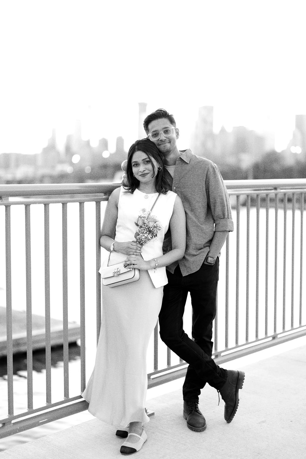 Black and white photo of a smiling couple posing on a bridge with a city skyline backdrop, woman holding flowers.