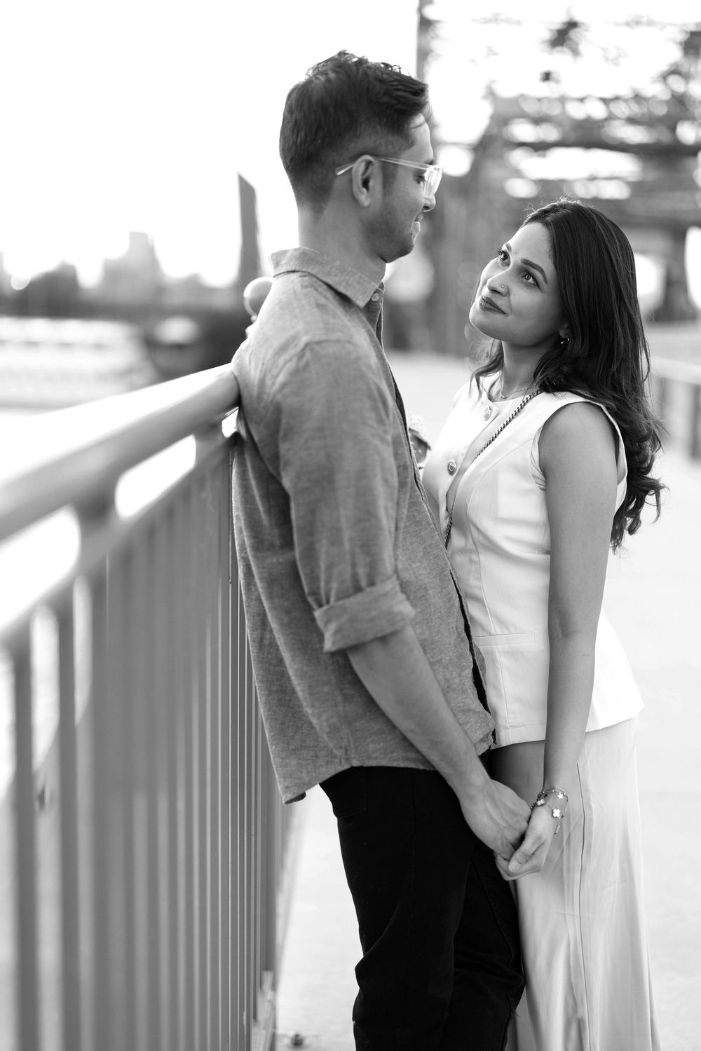 Black and white photo of a romantic couple holding hands and gazing at each other on a bridge.
