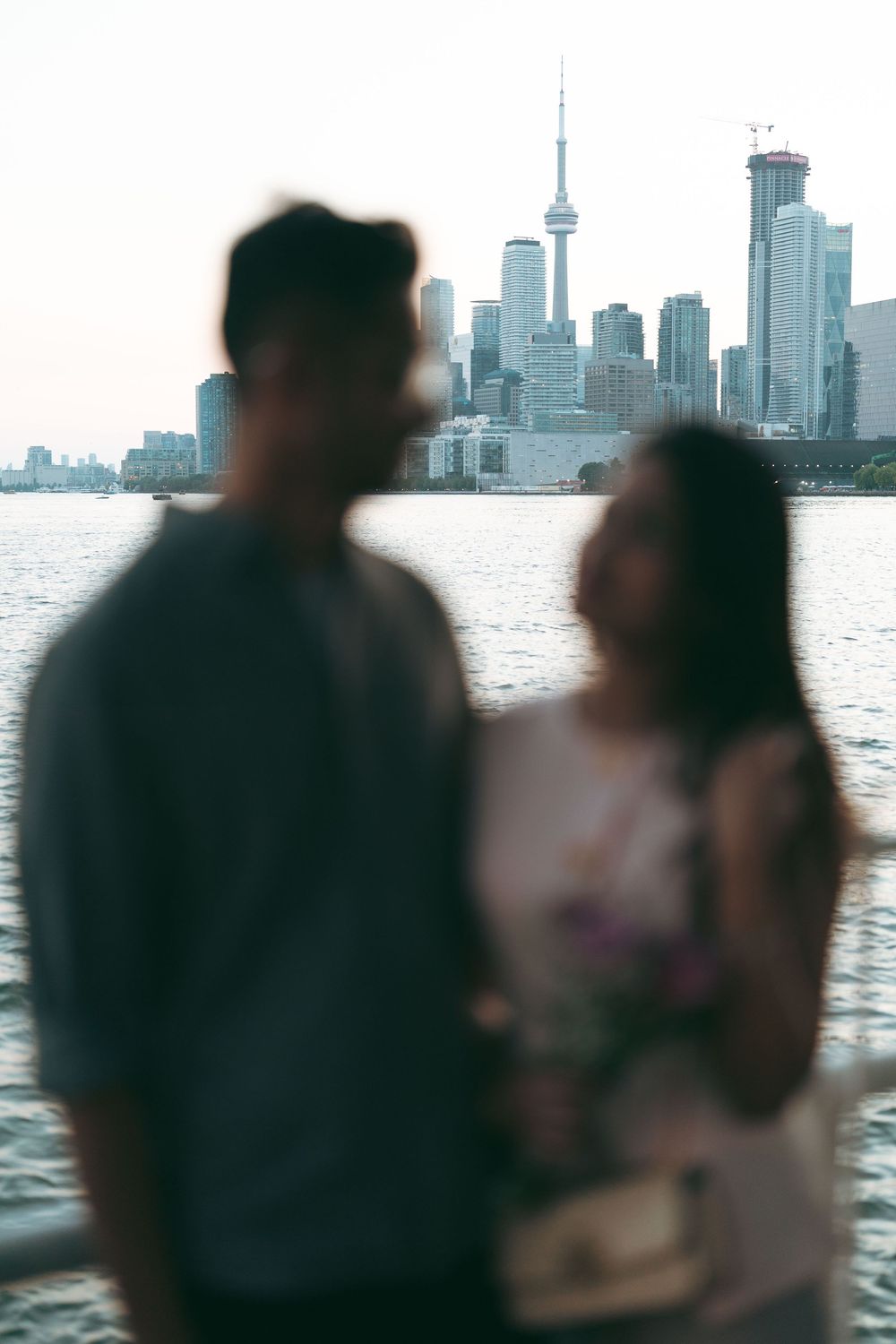 Couple standing together with the Toronto skyline and CN Tower visible across the water in the background.