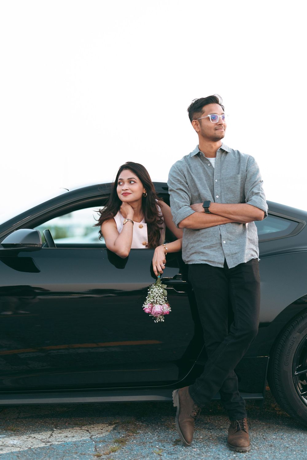 A young couple poses by a black car, woman leaning out window holding flowers, man standing with arms crossed wearing sunglasses.