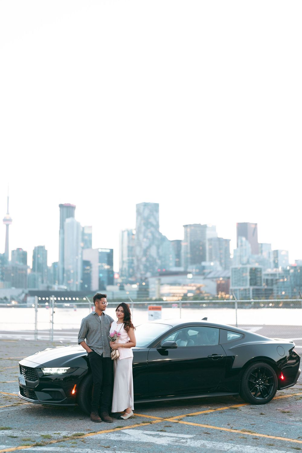 Couple posing beside a black sports car on a rooftop parking lot with Toronto city skyline in the background.