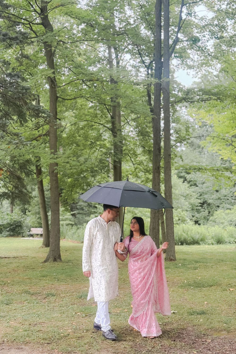 Indian couple in traditional attire sharing umbrella in lush green park, man in kurta, woman in pink saree.