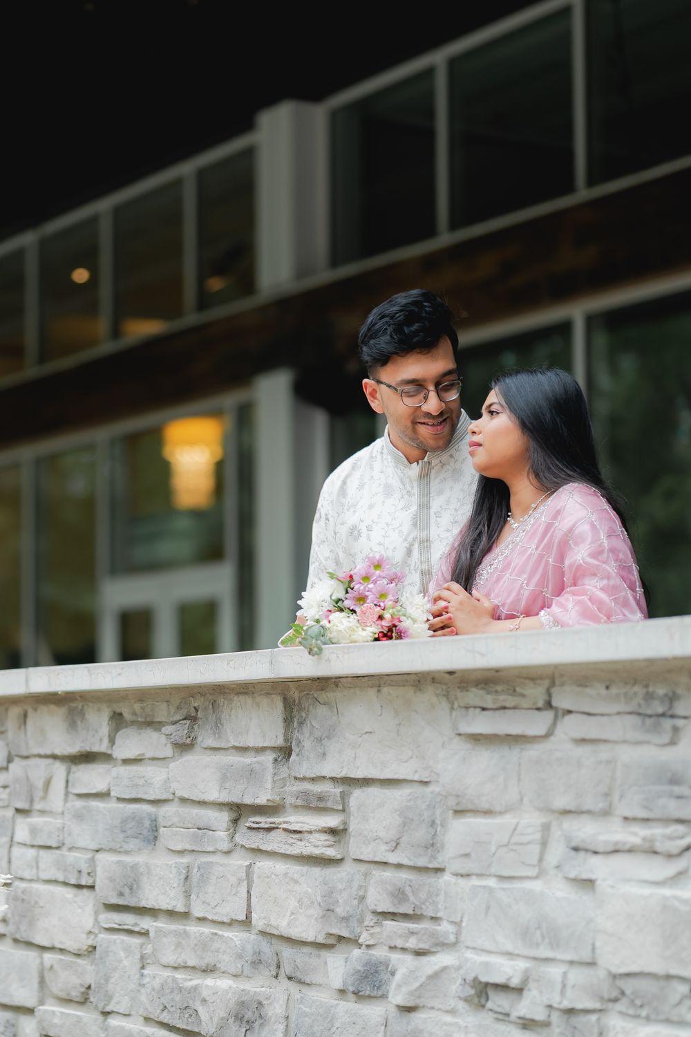 A couple in traditional South Asian attire stands together on a stone balcony, sharing a tender moment with flowers nearby.