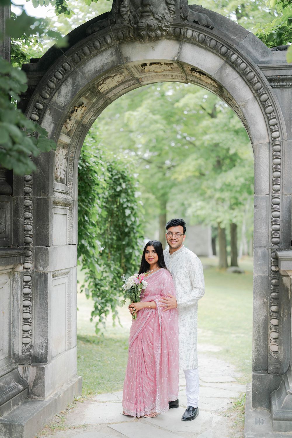 Couple in traditional South Asian attire posing under ornate stone arch in lush garden setting.