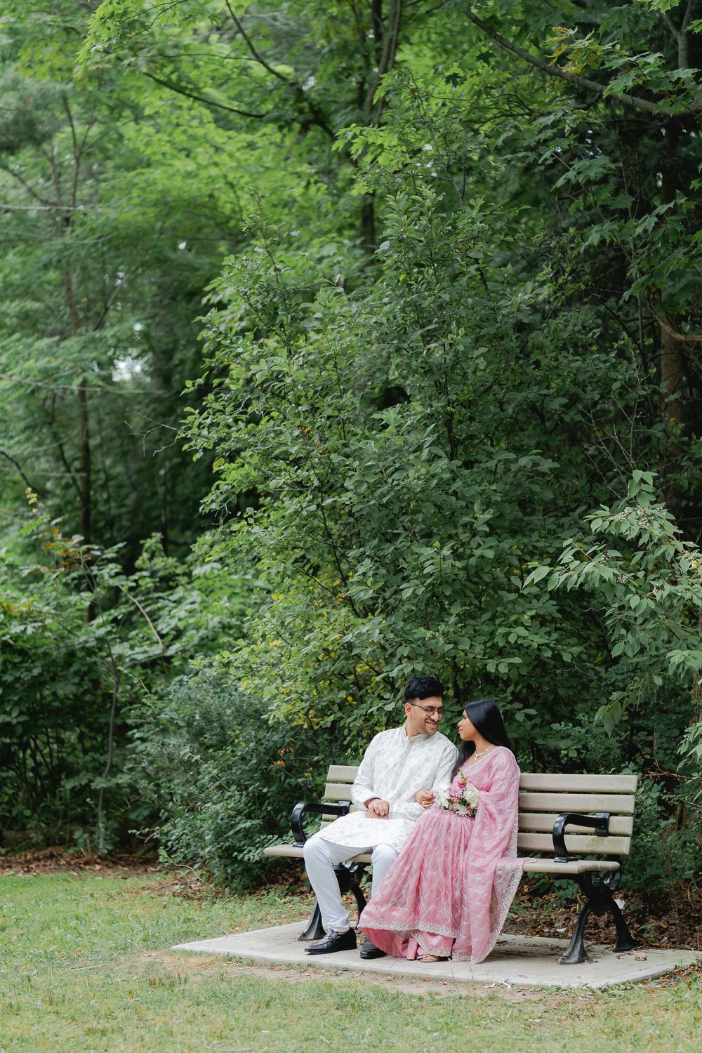 Couple in traditional South Asian attire sitting on a park bench surrounded by lush green trees.