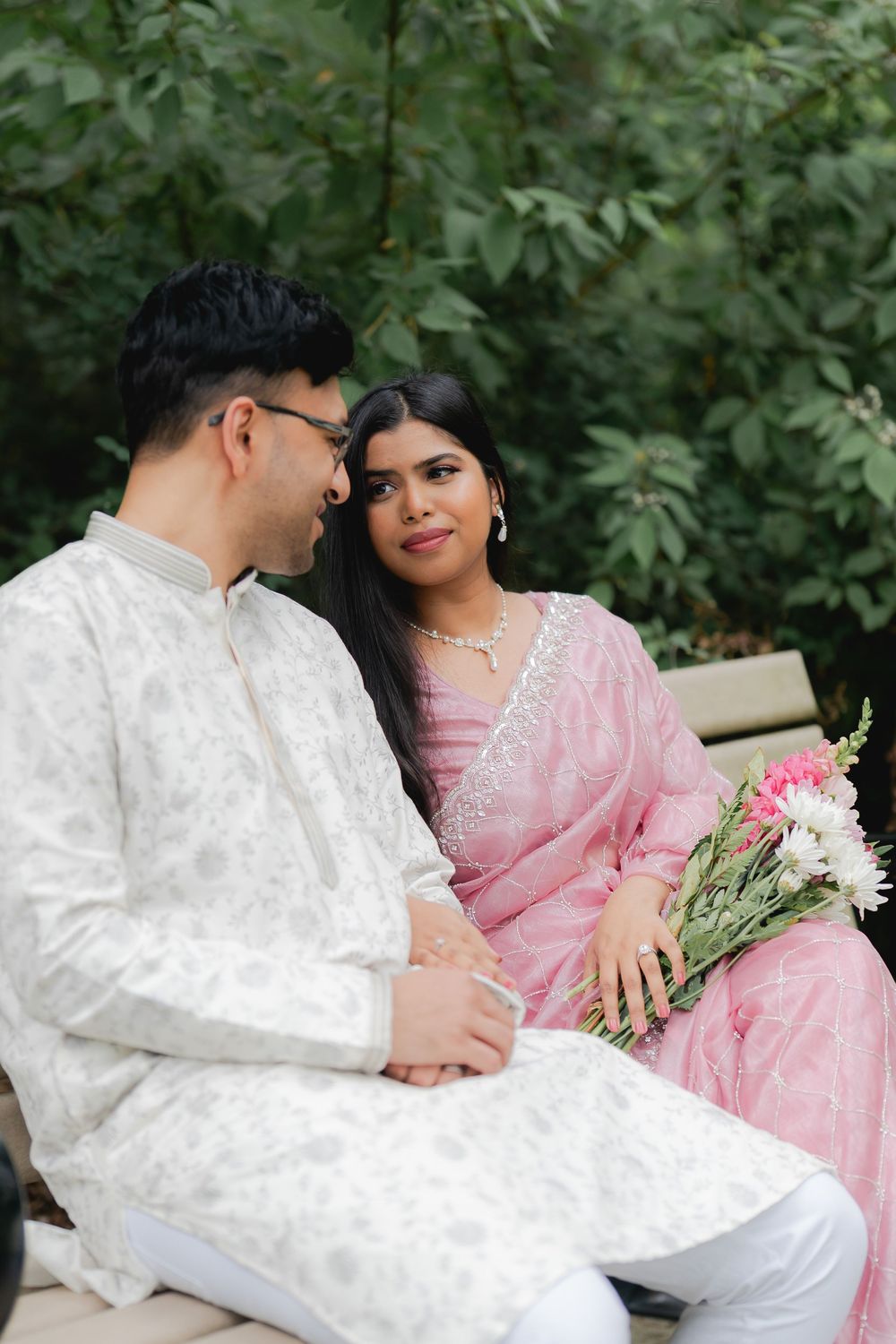 A couple in traditional South Asian attire sits outdoors, the woman in pink holding flowers, smiling at the man in white kurta.
