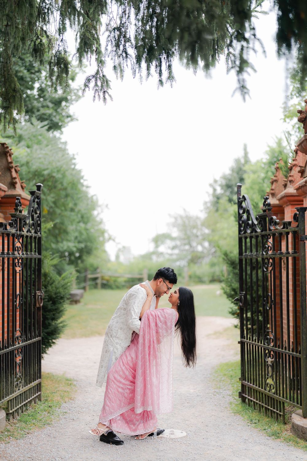 Couple sharing a romantic kiss in traditional South Asian wedding attire at ornate iron garden gates.
