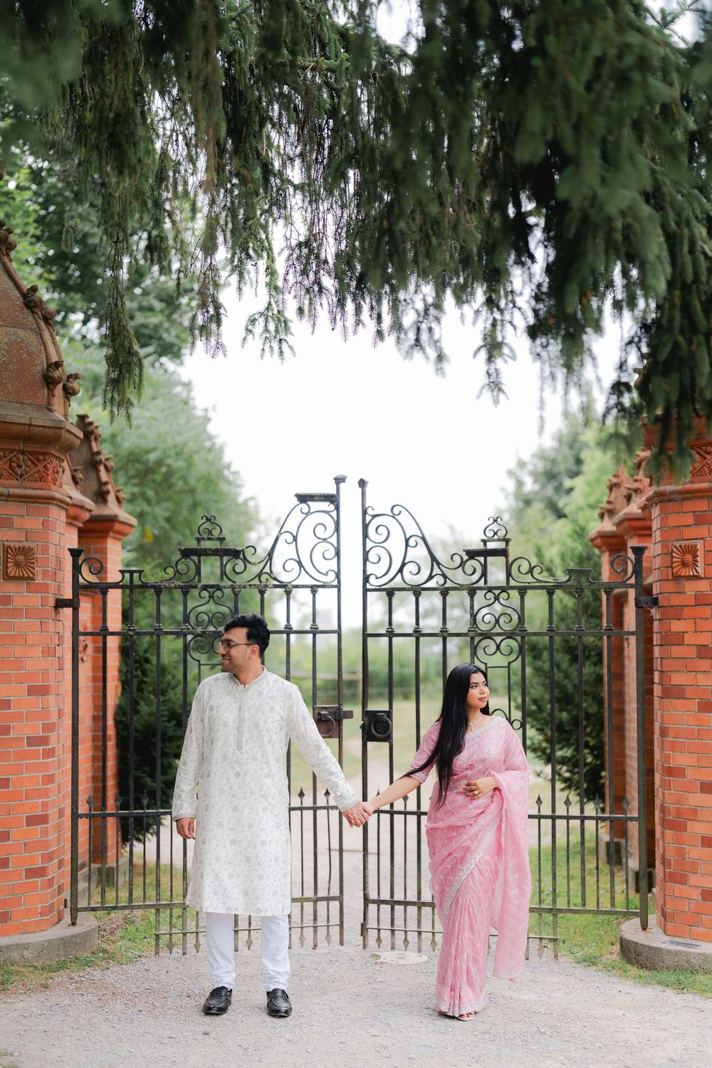 Indian couple in traditional attire holding hands by ornate iron gates with brick pillars and lush green trees.