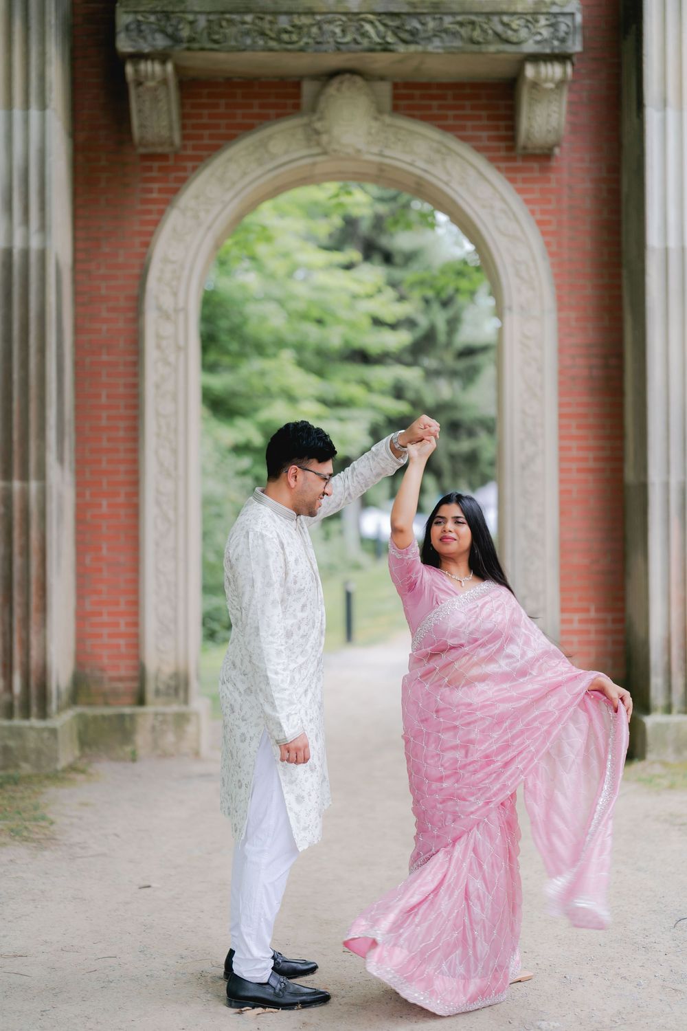 Couple in traditional South Asian attire dancing under a brick archway, man in white kurta, woman in pink saree.