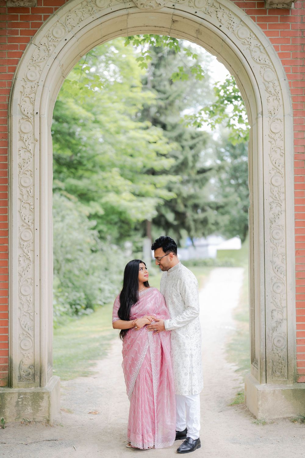 Couple in traditional South Asian attire embracing under ornate stone arch surrounded by lush green trees.