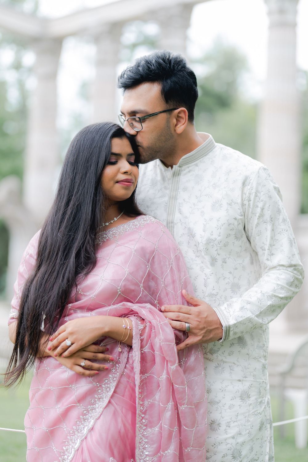 A couple in traditional South Asian attire, man kissing woman's temple while embracing outdoors near columns.