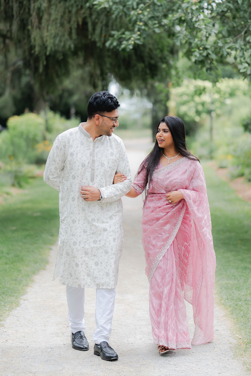 Couple in traditional South Asian attire walking in a lush garden, man in white kurta, woman in pink saree.