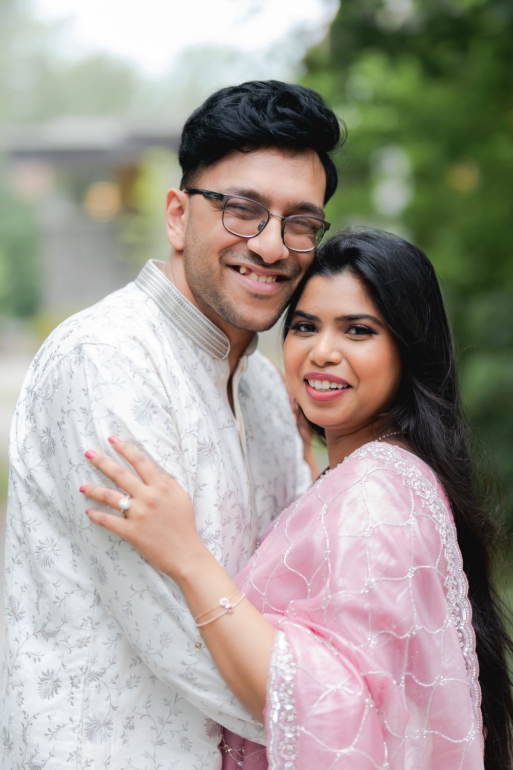 A smiling South Asian couple in traditional attire embrace outdoors, man in white kurta with glasses, woman in pink dupatta.