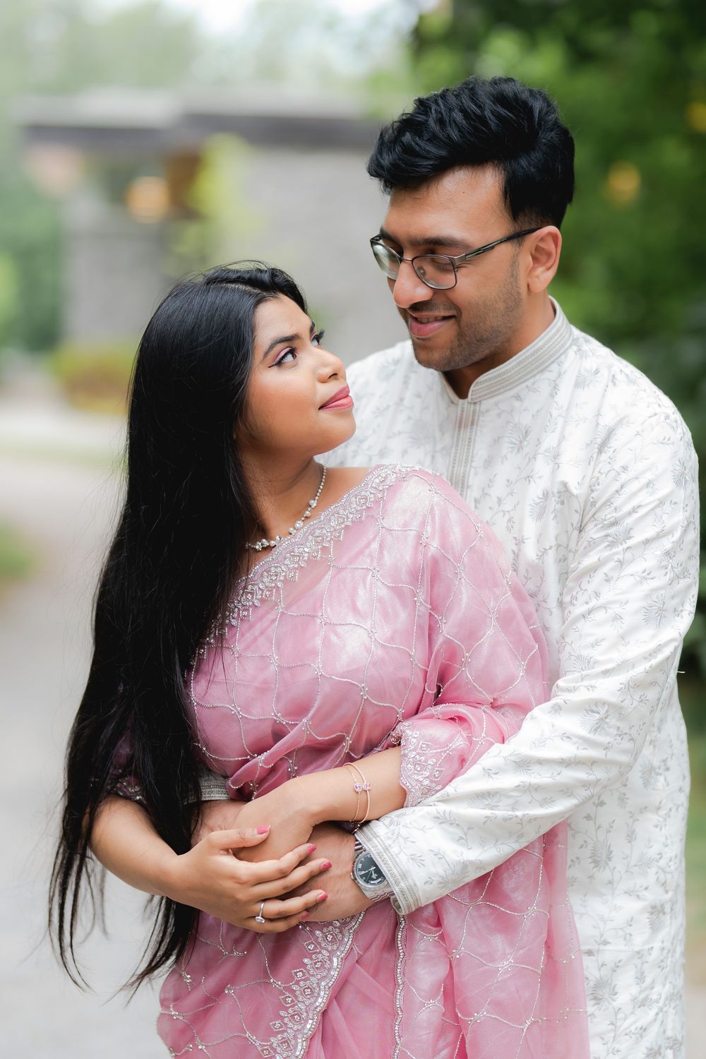 Couple in traditional South Asian attire, woman in pink saree and man in white kurta, sharing a romantic outdoor moment.