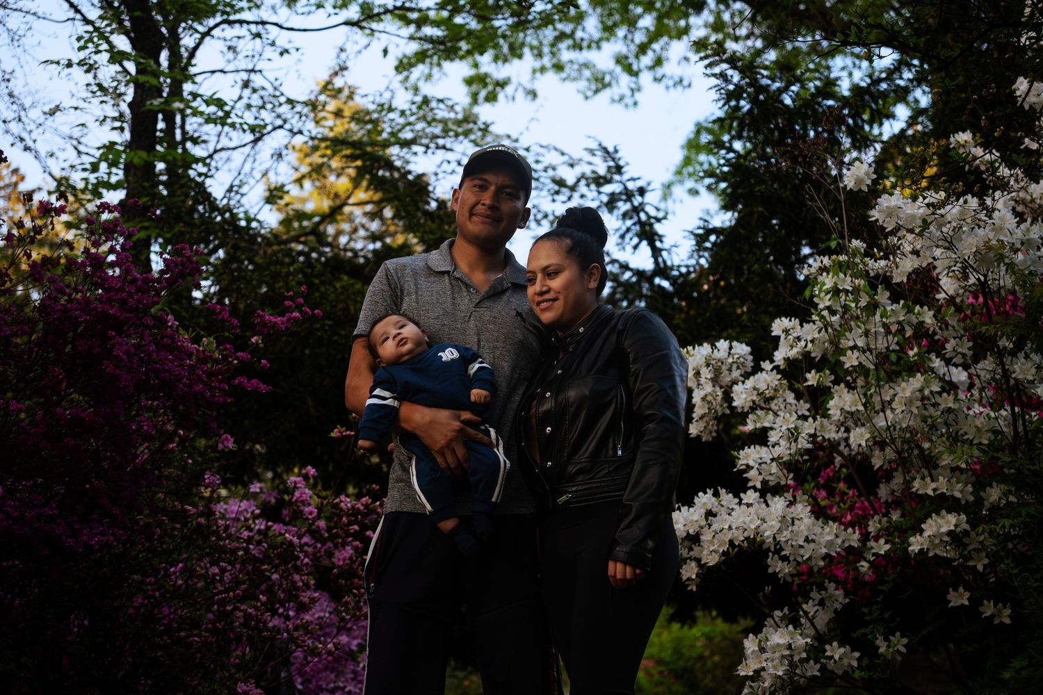 Parents holding infant baby posing outdoors among colorful blooming garden flowers.
