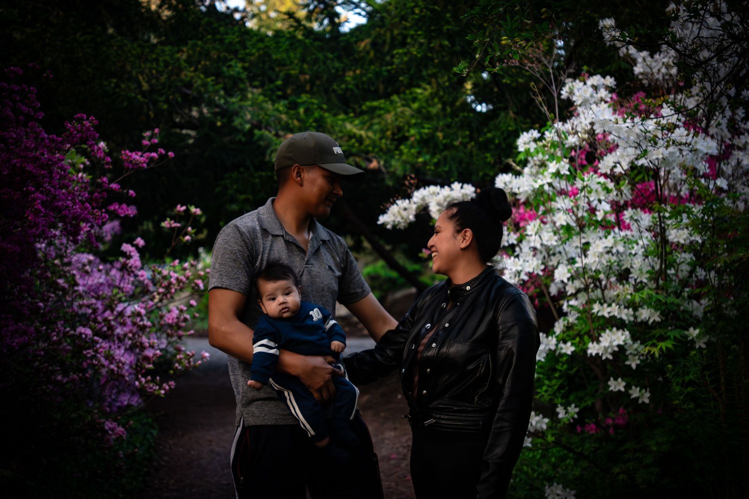 Family with baby posing in a garden surrounded by blooming pink and white flowers.