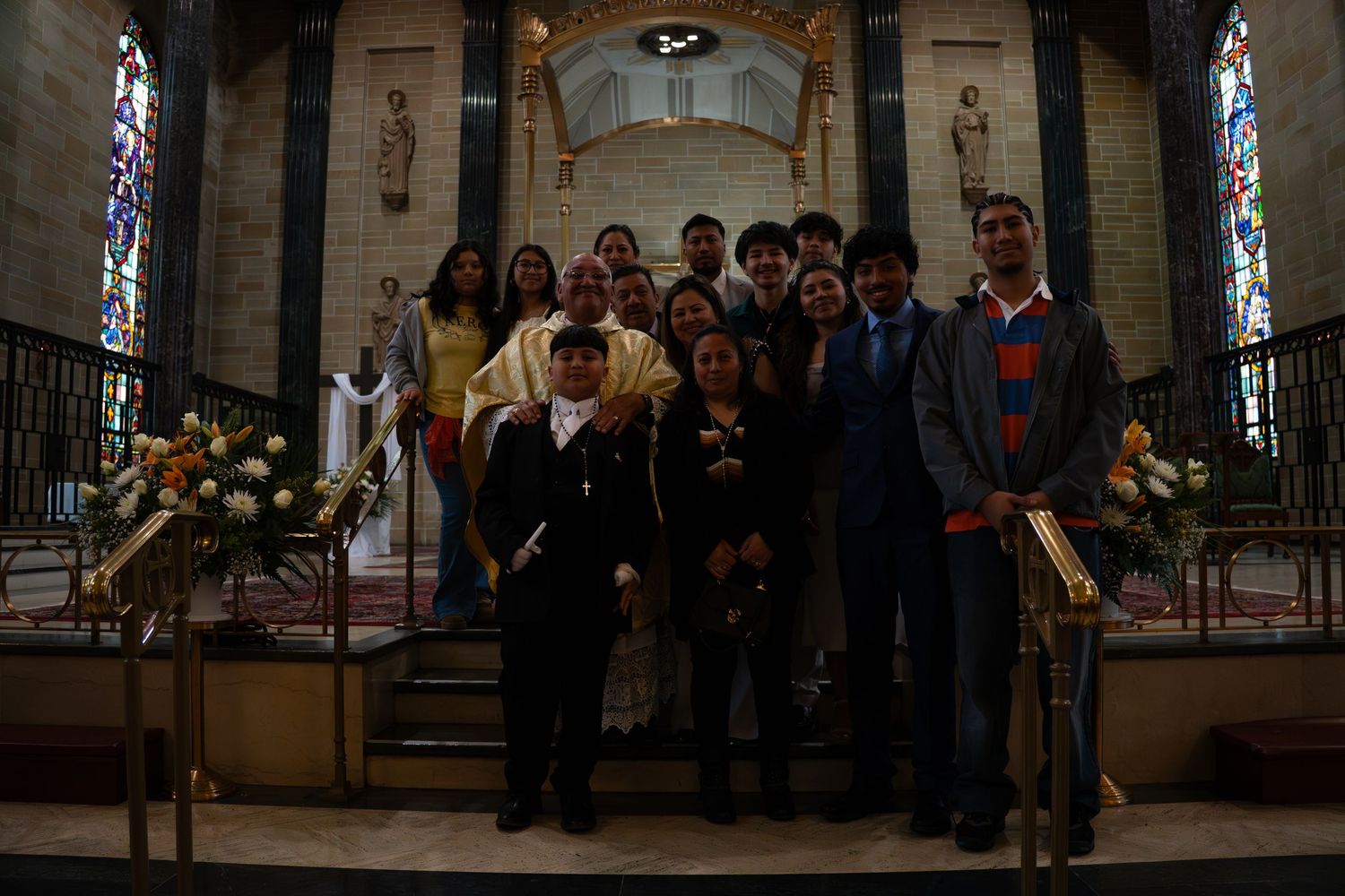 Group photo of people posing inside a church near the altar with stained glass windows and floral arrangements.