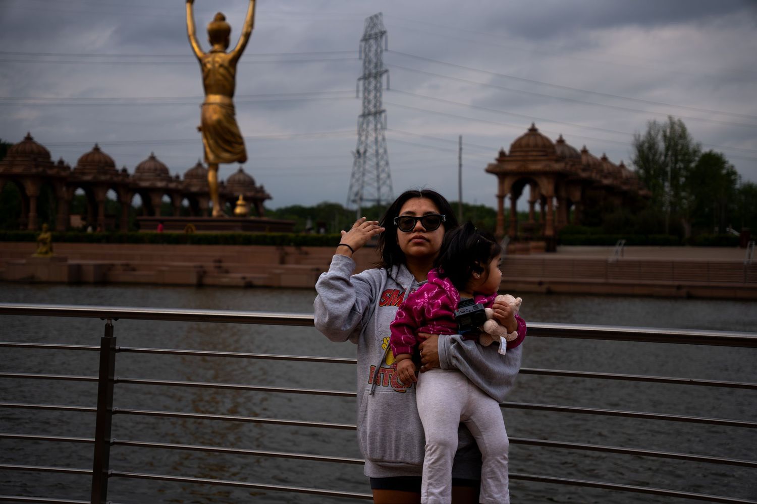 Woman holding child near a golden statue and temple complex under cloudy skies.