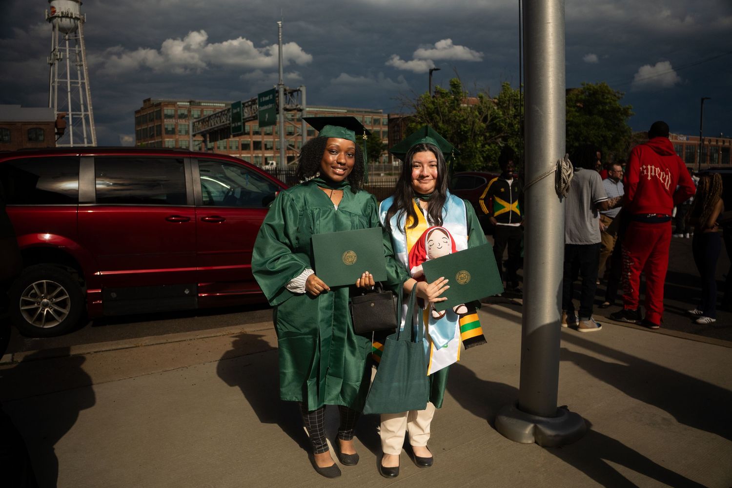 Two graduates in green caps and gowns holding diplomas, posing outdoors under dramatic cloudy sky.