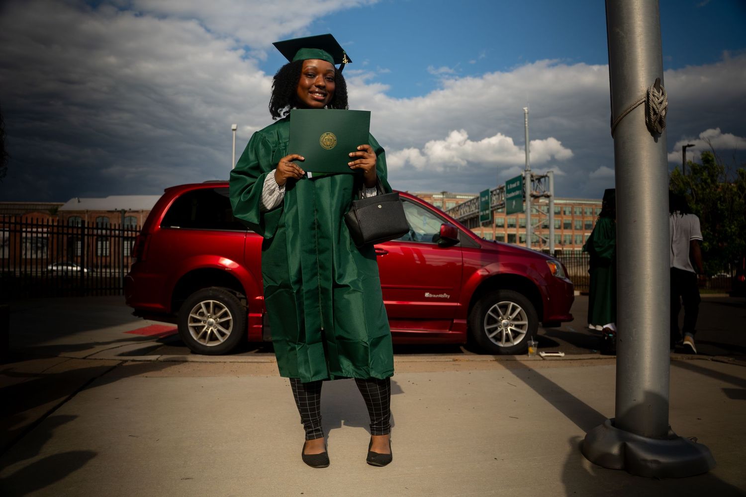 Female graduate in green cap and gown proudly holding diploma outdoors with cloudy sky background.