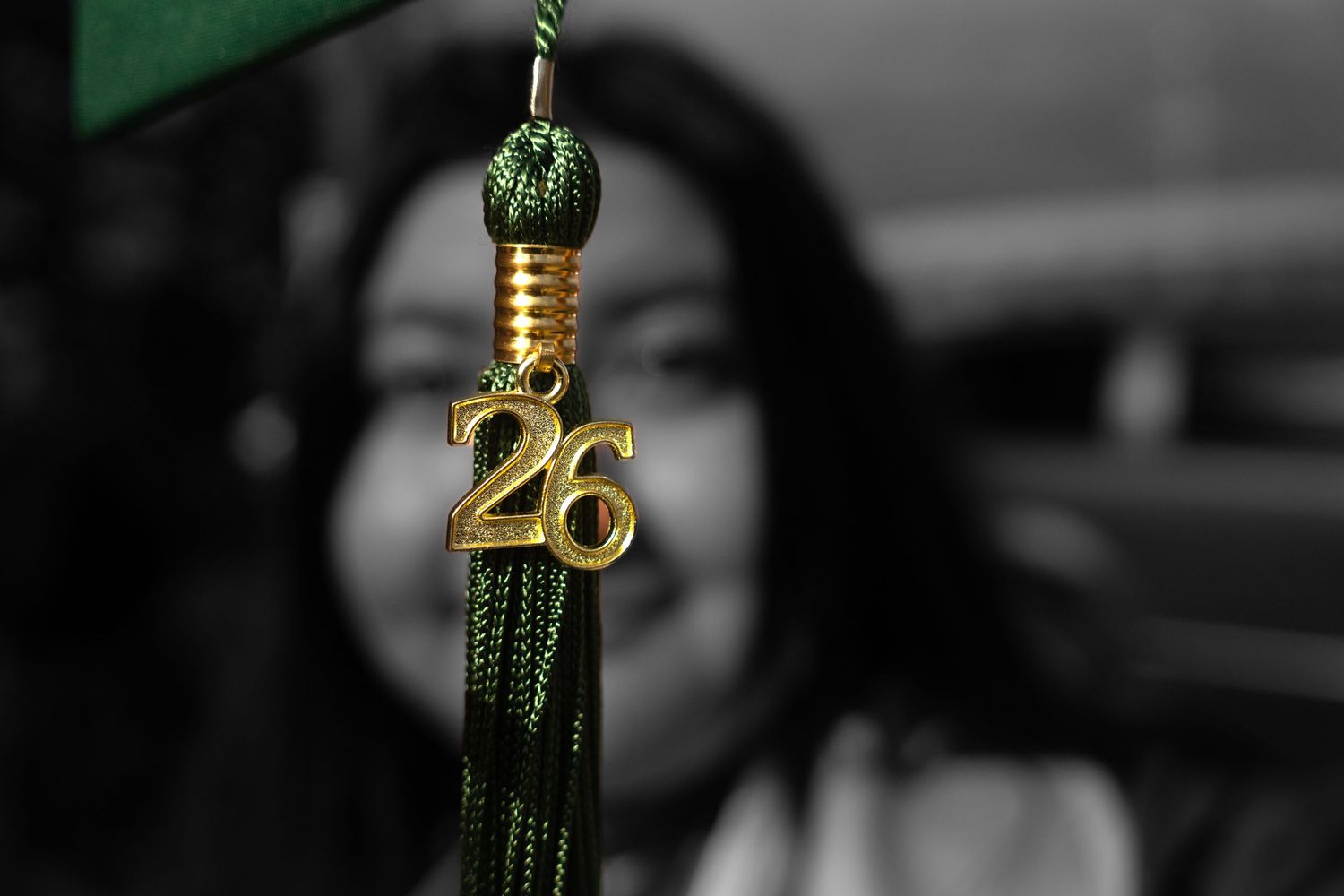 Close-up of green graduation tassel with gold number 26 charm, smiling graduate blurred in background.