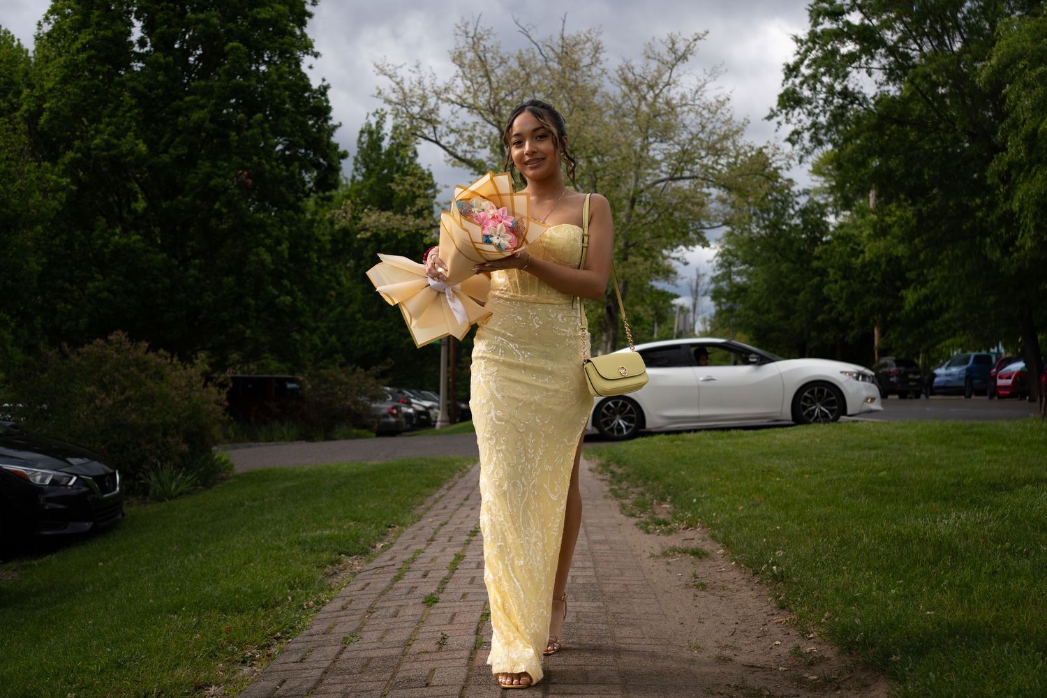 Young woman in elegant yellow gown walking on pathway holding colorful flower bouquet outdoors.
