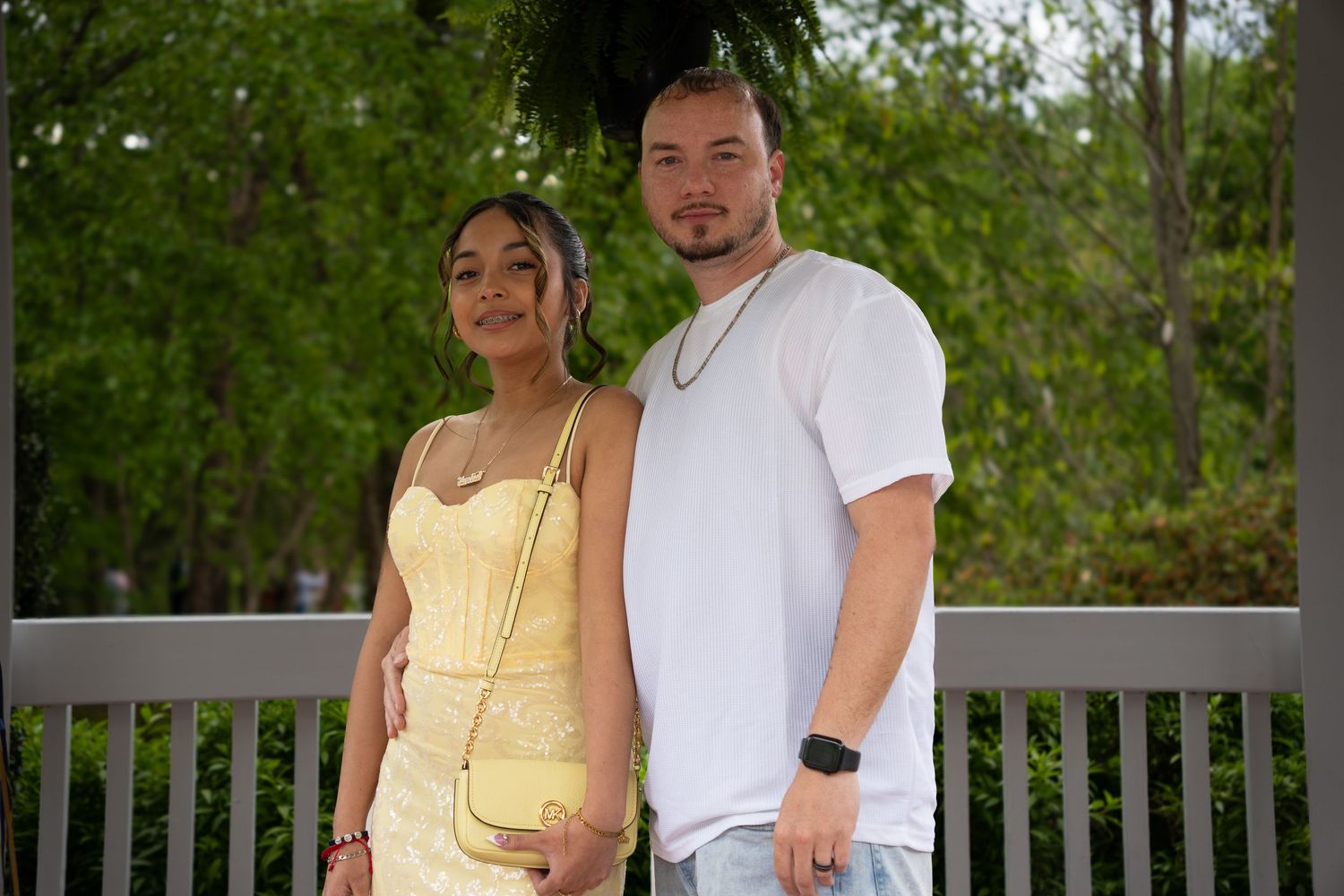Father and Daughter posing together outdoors, woman in yellow dress holding small purse, man in white t-shirt.