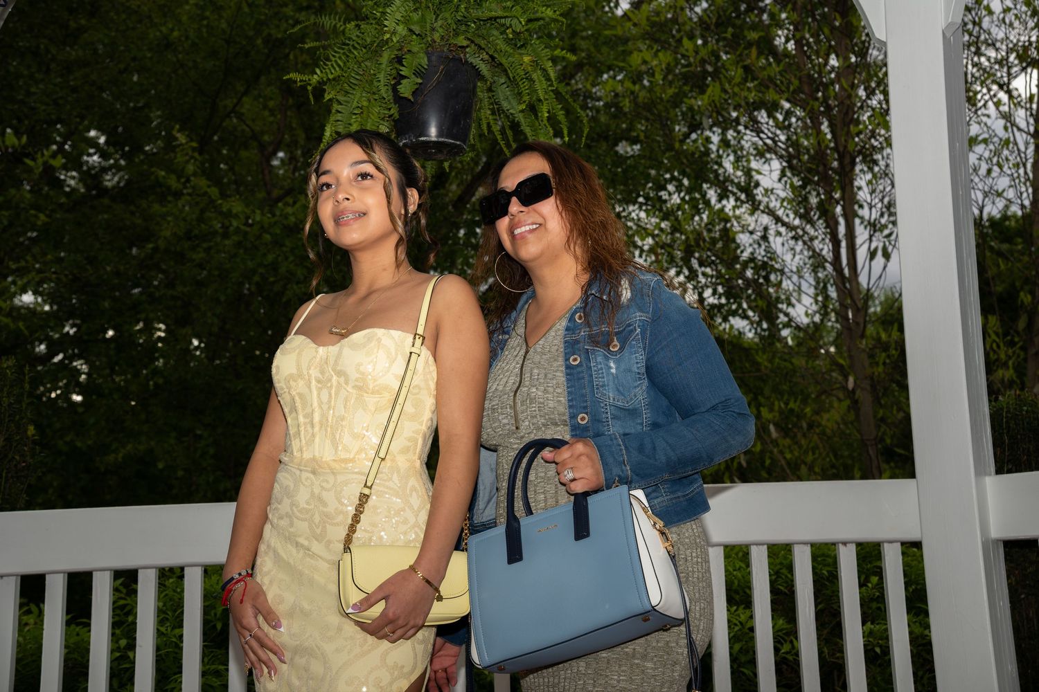 Mother and Daughter posing together on white gazebo, one in yellow dress, other in denim jacket holding blue purse.